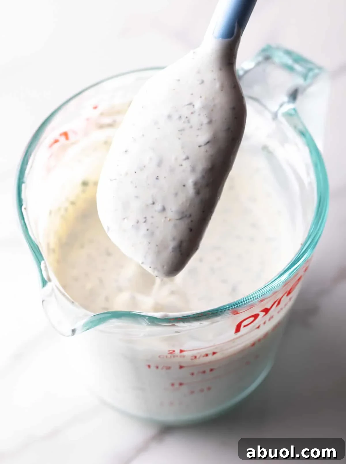 A spatula holding freshly made homemade buttermilk ranch dressing above a glass measuring pitcher filled with the dressing.