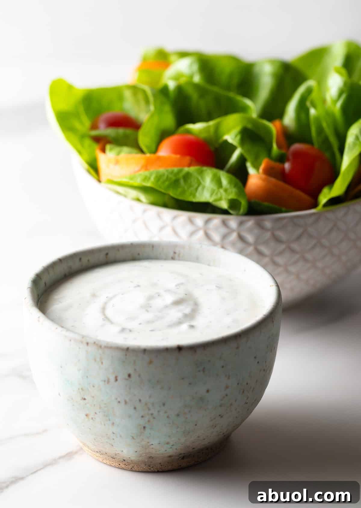 A white bowl filled with creamy homemade ranch dressing next to another bowl filled with salad.