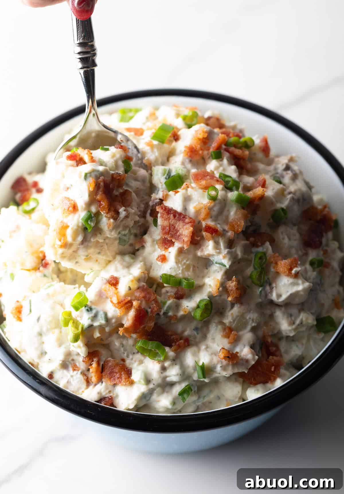 Bacon ranch potato salad is being mixed with a metal spoon in a bowl.
