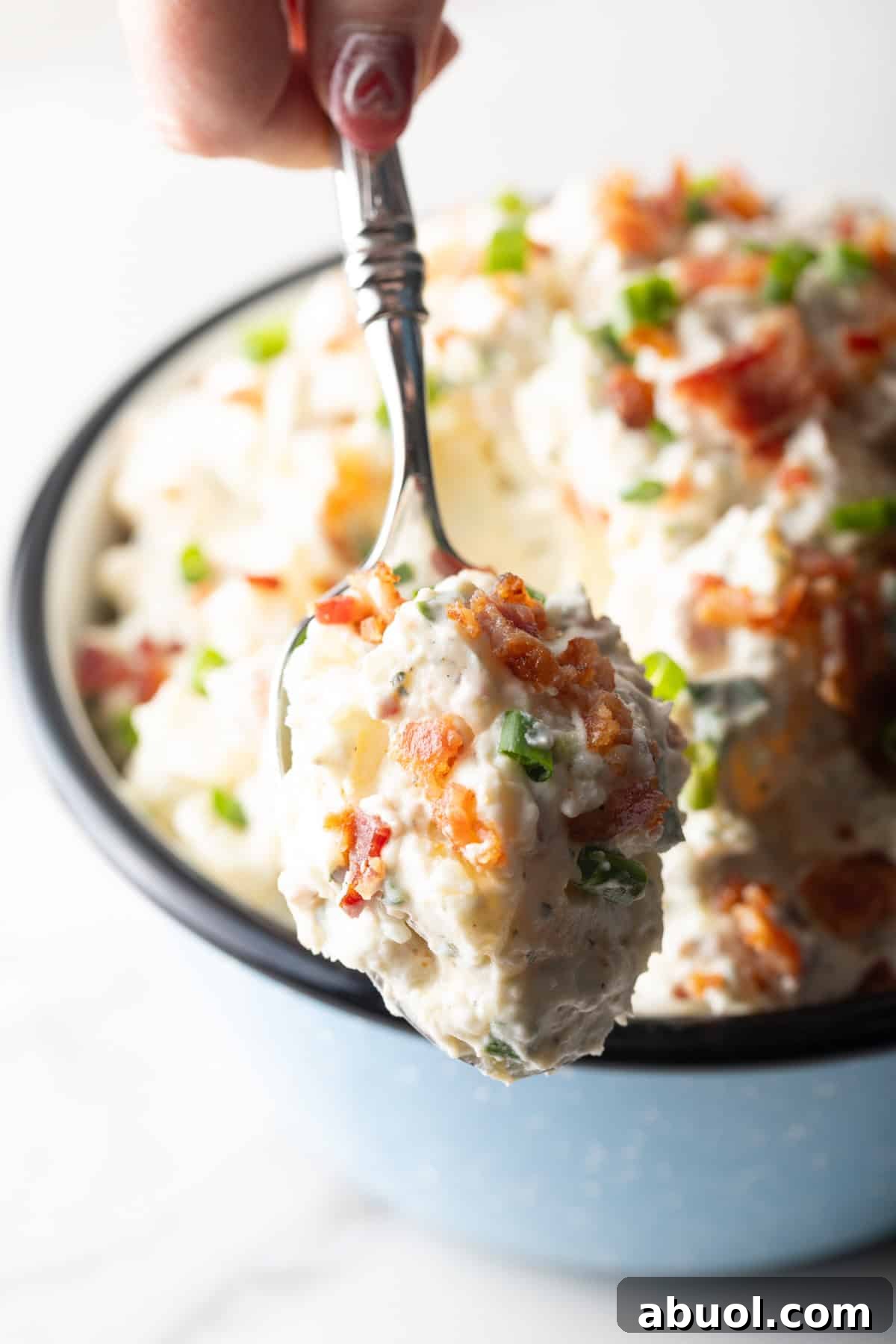 A spoonful of ranch potato salad with bacon is being held above a bowl of the potato salad.