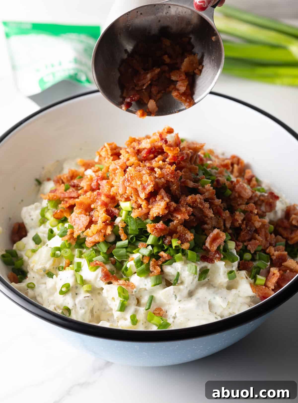 Chopped bacon is being poured over a bowl of bacon ranch potato salad, ready to be mixed in with the other ingredients on top.