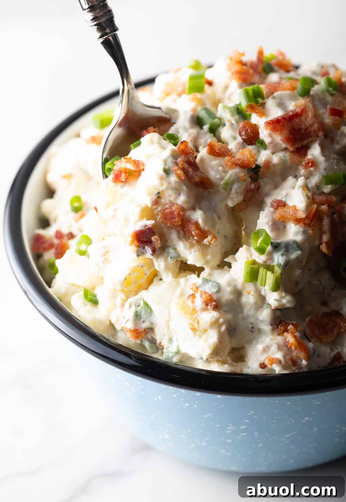Bacon ranch potato salad is being mixed with a metal spoon in a bowl.
