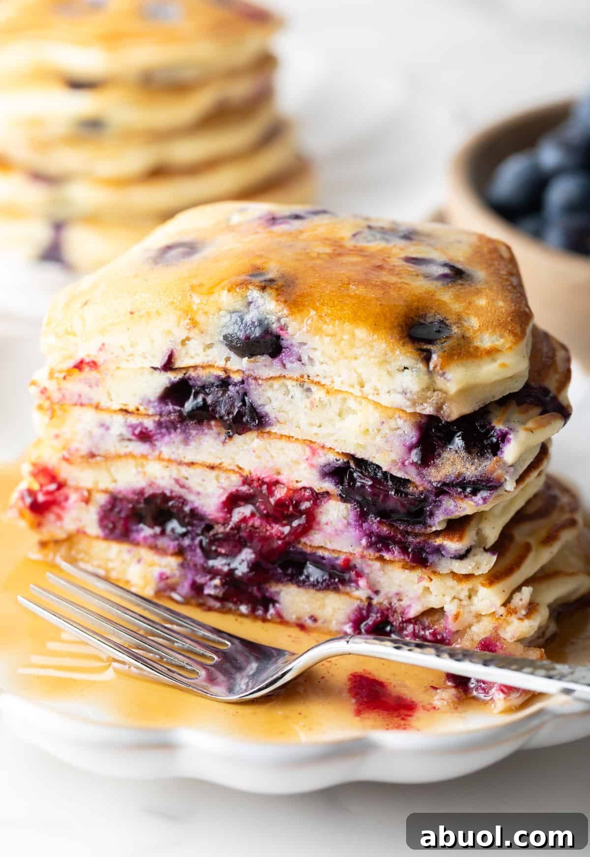 Stack of several blueberry pancakes on a plate, close up view of the pancakes cut in half to show the berries inside. There is a light puddle of syrup and fork on the plate.