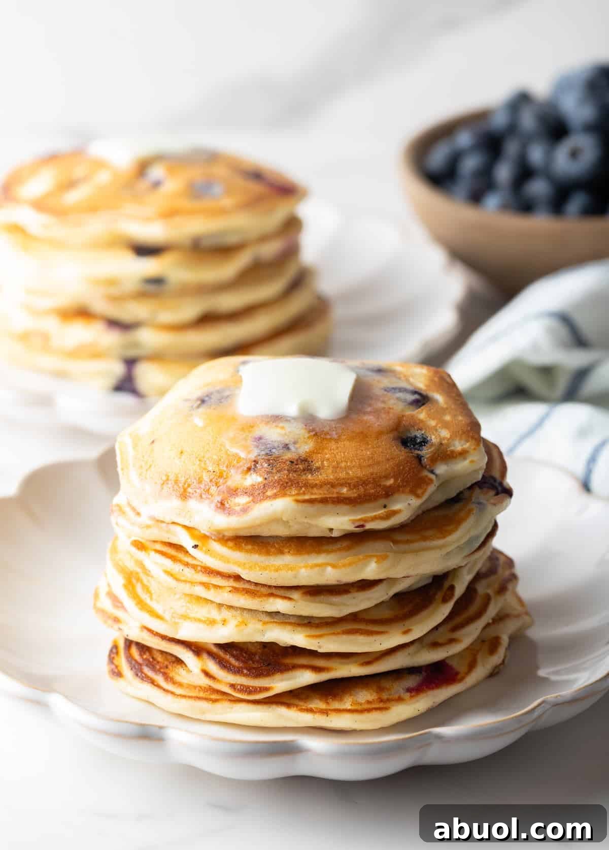 A large stack of blueberry pancakes in the foreground, another stack of pancakes in the background with a bowl of fresh berries. The stack closest to camera has a pat of butter on top.