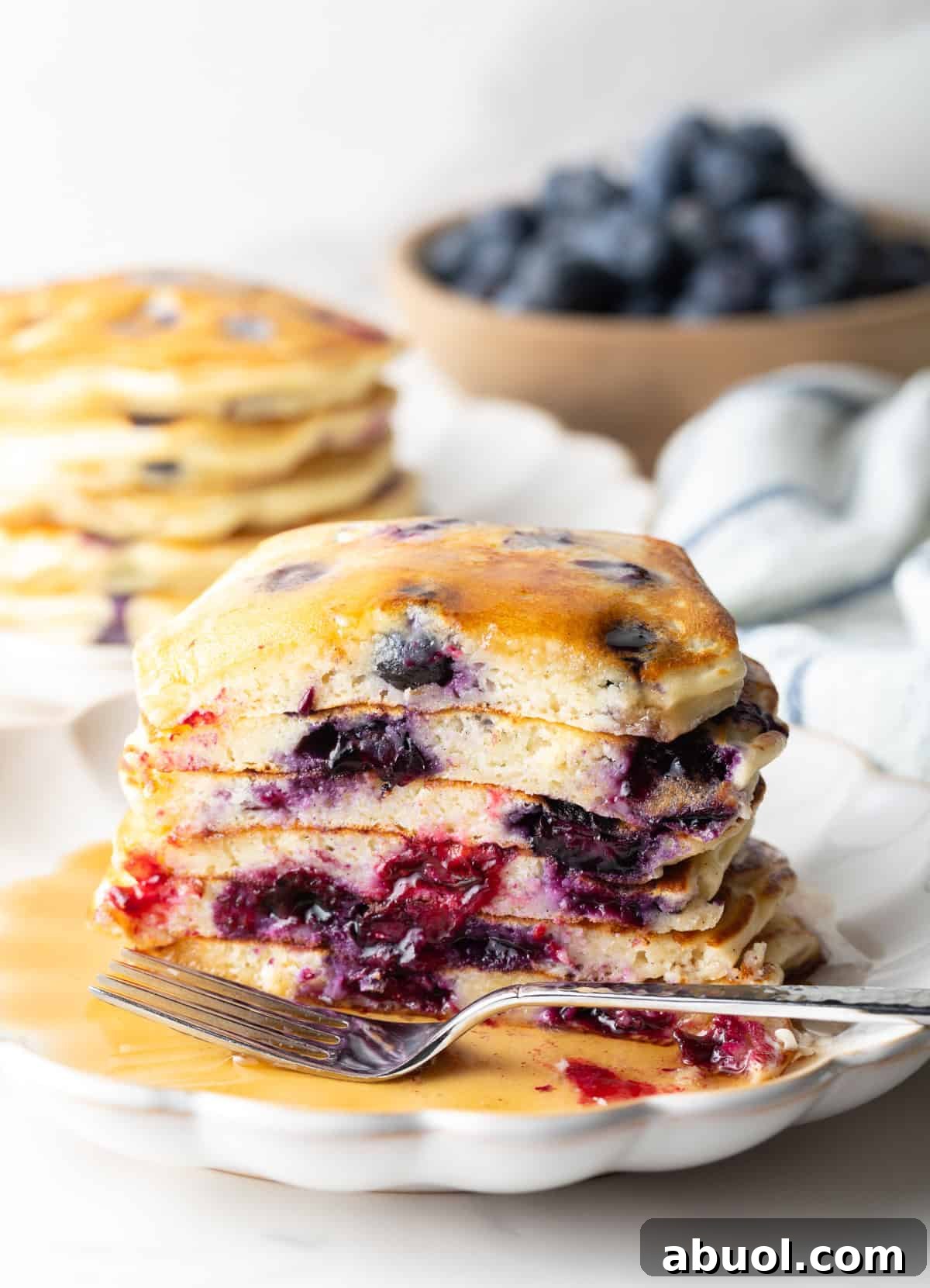 Stack of several blueberry pancakes on a plate, close up view of the pancakes cut in half to show the berries inside. There is a light puddle of syrup and fork on the plate.