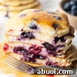 Stack of several blueberry pancakes on a plate, close up view of the pancakes cut in half to show the berries inside. There is a light puddle of syrup and fork on the plate.