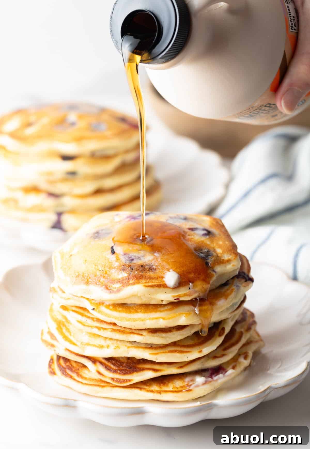 A large stack of pancakes on a white plate, being coated in a drizzle of syrup.