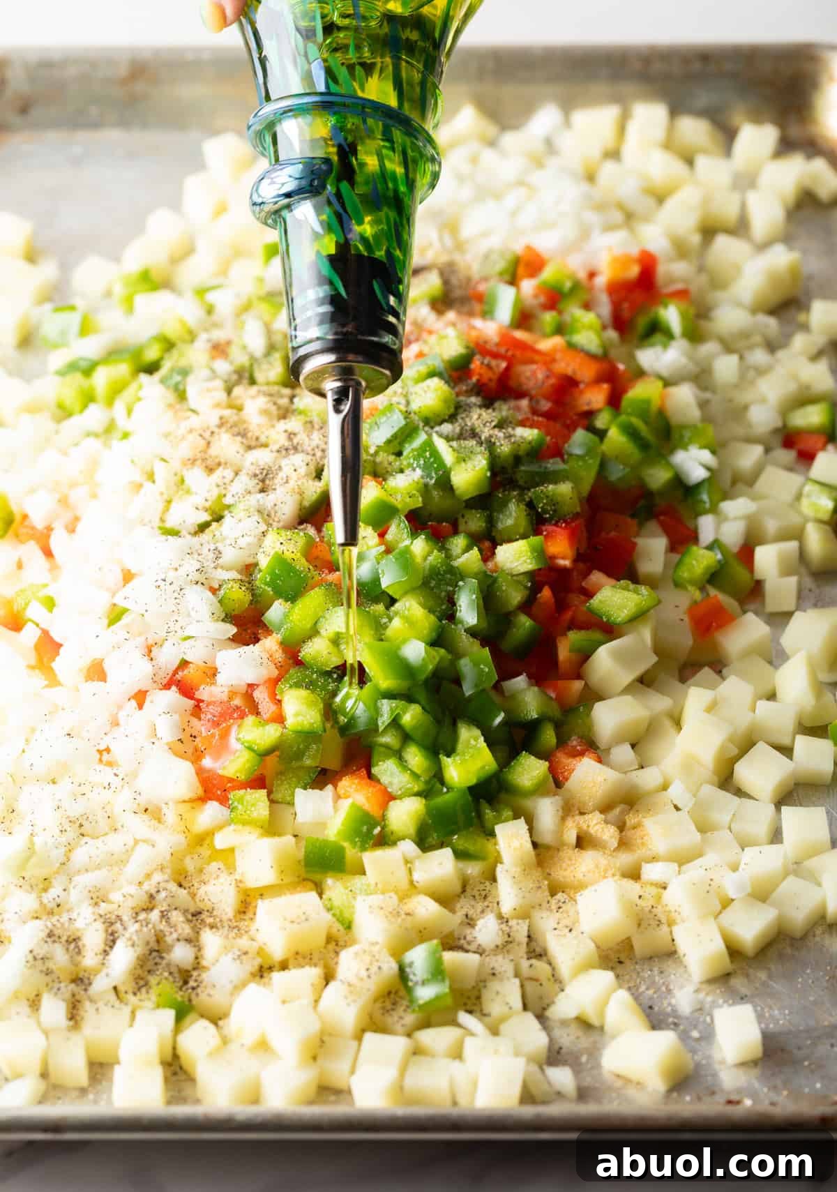 Olive oil is being drizzled over diced potatoes, bell peppers, and onion on a baking sheet.