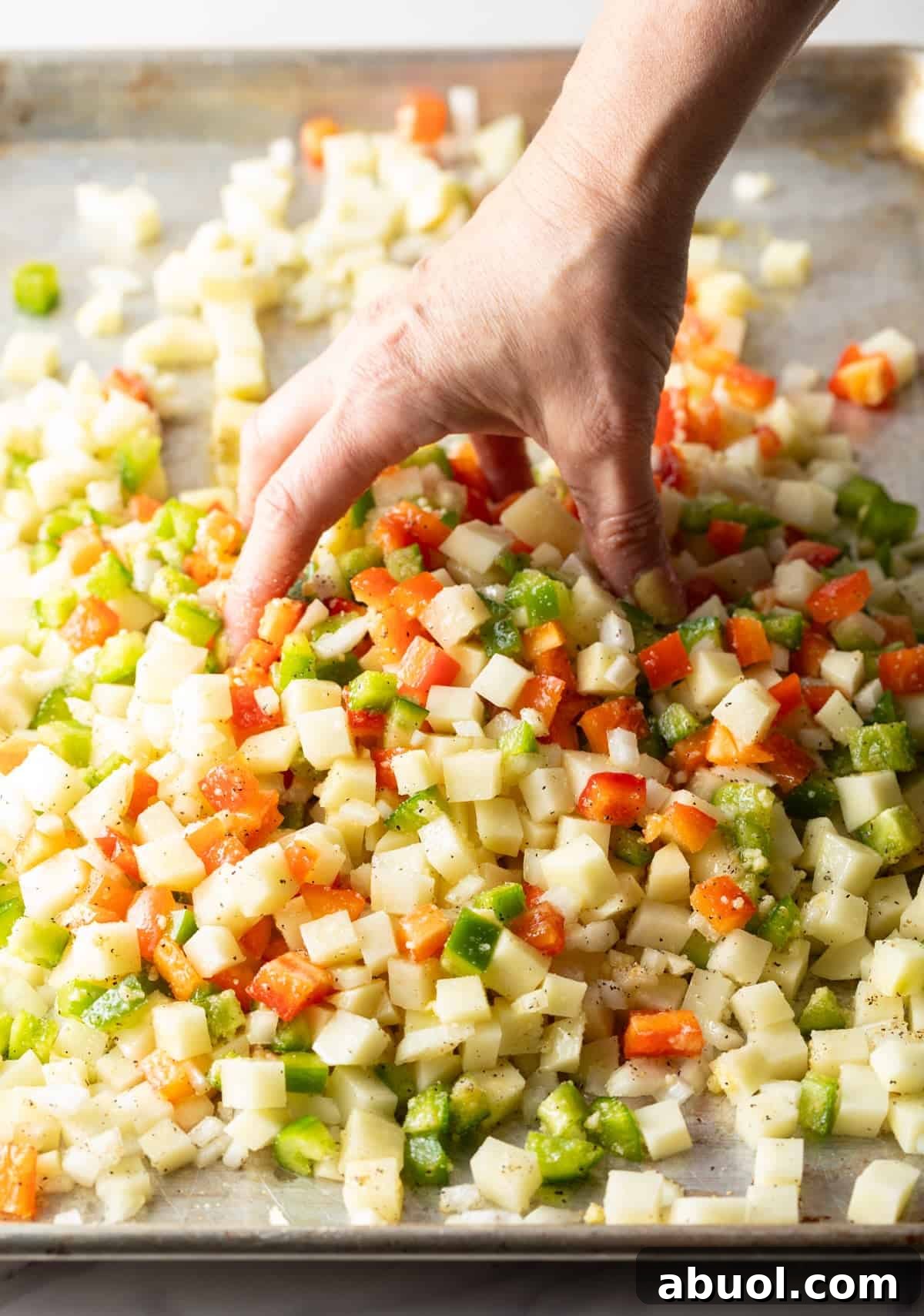 A hand is tossing the ingredients for a potatoes O'brien recipe with oil and seasonings on a baking sheet.