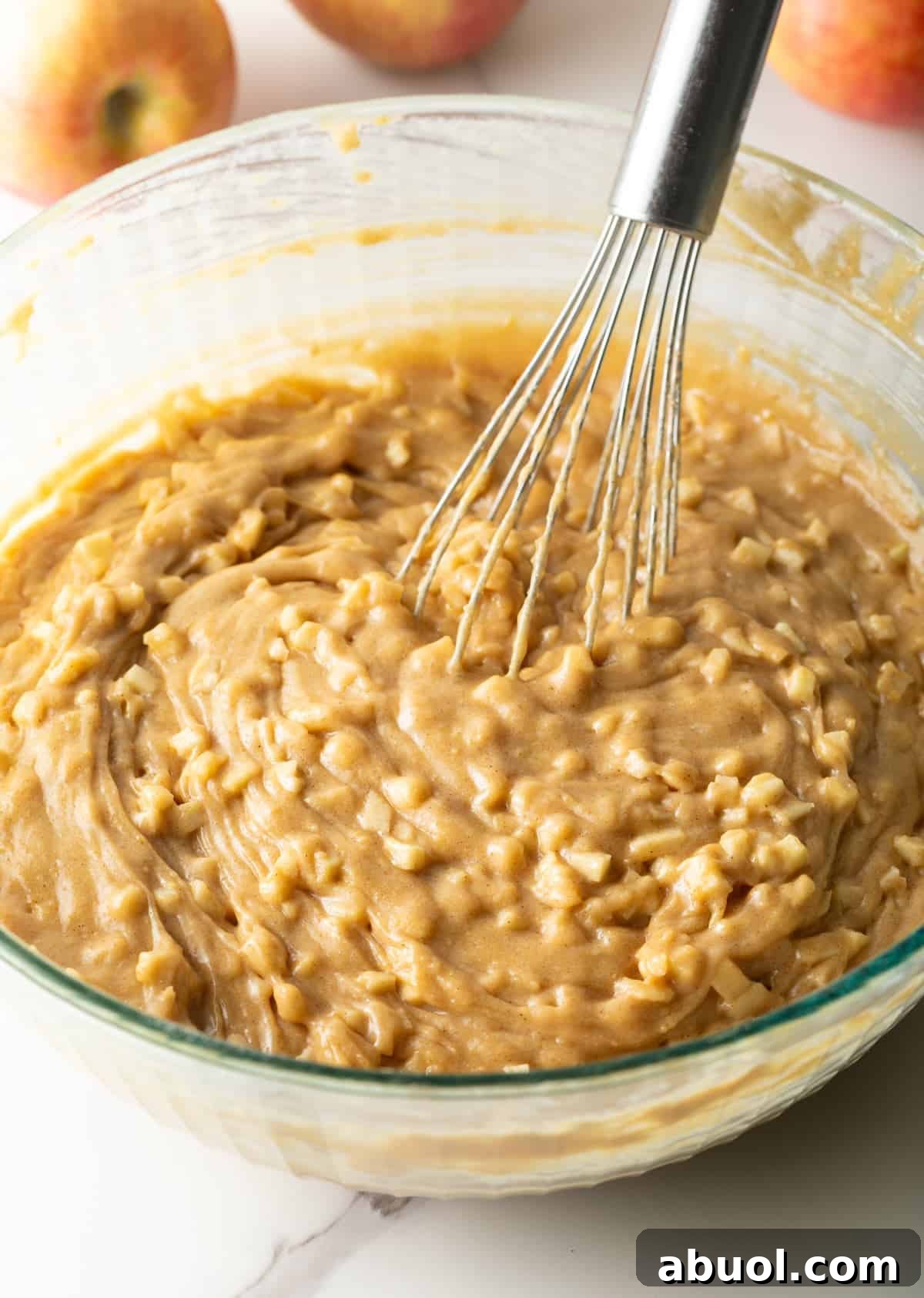 Diced apples have been added to an apple cinnamon bread mixture that is being stirred by a whisk.
